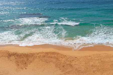 Golden sand stretches along the beach as gentle waves lap the shore under a clear blue sky. Sunbathers relax on towels in the warmth.の写真素材