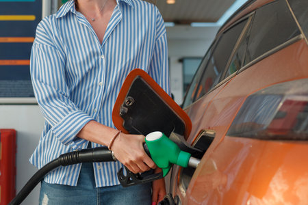 A person holds a fuel nozzle, refueling an orange vehicle at a gas station during the afternoon. The atmosphere is busy and practical.の写真素材