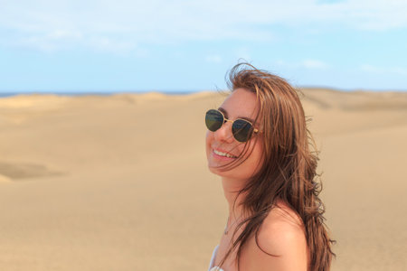 A joyful woman stands in the sandy dunes, wearing sunglasses and a breezy outfit, with wind-swept...