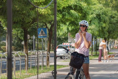 A woman wearing a helmet smiles while resting her bicycle beside a tree-lined walkway on a warm day,...