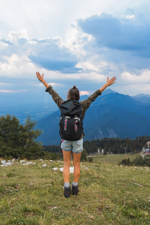A person with a backpack stands with arms raised at a mountain peak, enjoying the breathtaking...