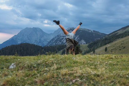 A person performs a handstand on a grassy hill as twilight approaches, with majestic mountain peaks...