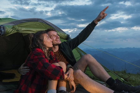 A couple sits together outside a tent, pointing at the clouds above while enjoying a serene moment...