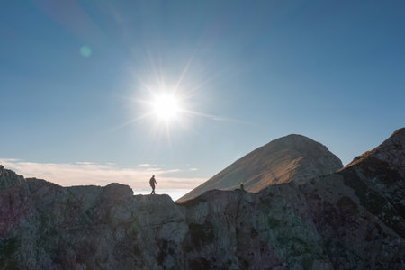 A lone hiker traverses a rugged mountain ridge as the sun shines brightly overhead, illuminating the...