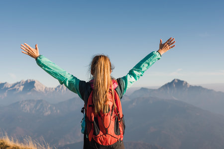 A woman with long hair raises her arms in joy atop a mountain, surrounded by stunning peaks under a...