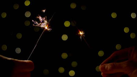 Two hands hold sparklers, creating bright sparks against a dark background filled with soft glowing...