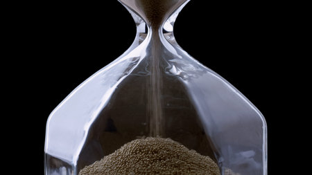 Fine sand flows from the top to the bottom of a clear hourglass, marking the passage of time.