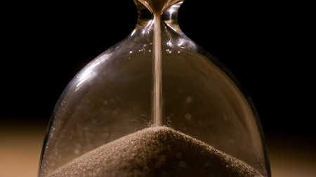 Fine sand trickles from the top chamber to the bottom of a clear glass hourglass against a dark...