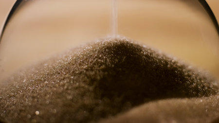 Fine grains of sand cascade gently inside a glass hourglass, marking the passage of time in a serene...