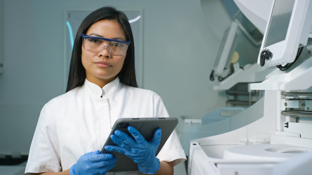 A female lab technician in protective gear examines data on a tablet while surrounded by laboratory...