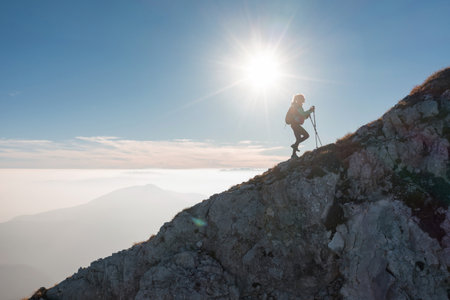 A lone woman hiker ascends a rocky mountain trail as the sun rises, casting a warm glow over the landscape.の写真素材