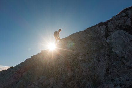 A person climbs a rocky slope as sunlight breaks through the mountains, creating a stunning silhouette.の写真素材