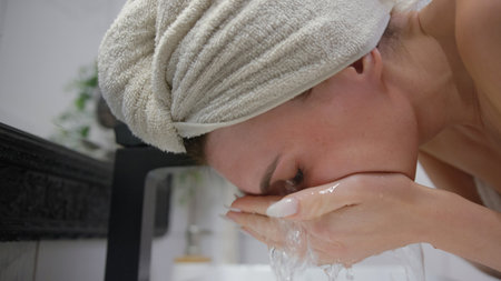 A woman with a towel on her head is washing her face at a bathroom sink, focusing on her skincare...