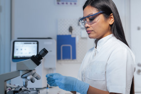 Focused female laboratory researcher observing slide through a microscope, dedicated to examining a...