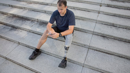 A young man with a prosthetic leg sits on stone steps, focused on his phone, enjoying a quiet moment outdoors.の写真素材