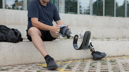 A person sits on a bench, carefully adjusting their prosthetic leg at a public park in daylight.
