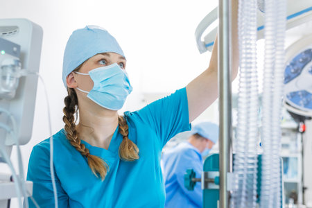 A healthcare worker in scrubs and a mask carefully adjusts medical equipment in a bright hospital room during the day.の写真素材