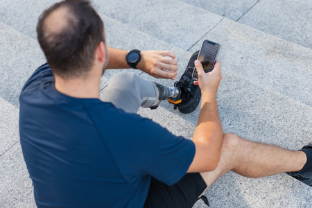 Man with disability, wearing a leg prosthesis, sitting on the street steps, looking at a wrist watch. Physical impairment and mobility concept.の写真素材