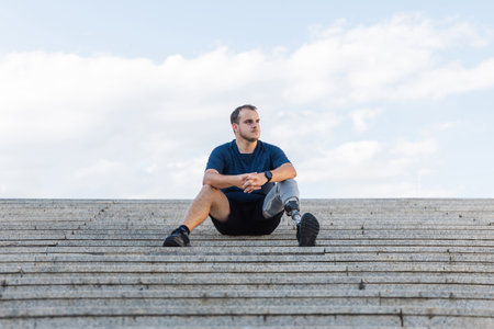 Portrait of a young confident man with disability, wearing a prosthetic leg, sitting outdoors, on...