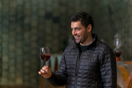 A man smiles while holding a glass of red wine in a cozy winery setting, surrounded by wooden...