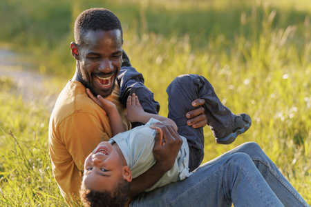 African American father sitting in a sunny grassy field, holding and gently roughhousing with his...