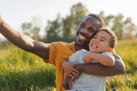 A father smiles joyfully while holding his young son in a grassy field during daylight, capturing a special moment selfie.の写真素材