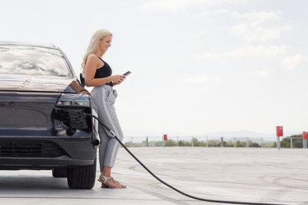 A woman stands by an electric car while charging it at a station. She uses her phone while waiting.