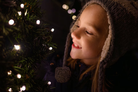 A child enjoys the glow of warm festive lights on a Christmas holiday tree, smiling happily during a...