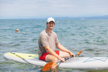 Young man paddles on a bright summer day while stand up paddleboarding in the sea. He enjoys the...
