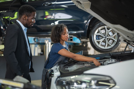 Female mechanic pointing at engine parts while holding a tablet displaying diagnostic data, explaining the repair to a male client.