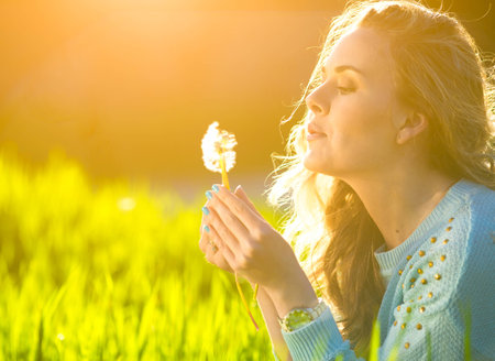 Beautiful young woman blowing dandelion. Trendy young girl at sunset with flowerの写真素材