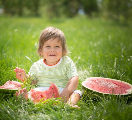 Happy child with big red slice of watermelon sitting on green grass. Healthy eating conceptの写真素材