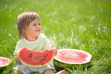 Happy child with big red slice of watermelon sitting on green grass. Healthy eating conceptの写真素材