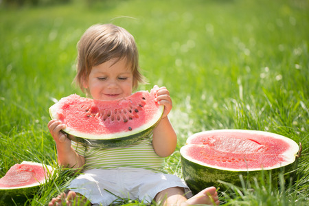 Happy child with big red slice of watermelon sitting on green grass. Healthy eating conceptの写真素材