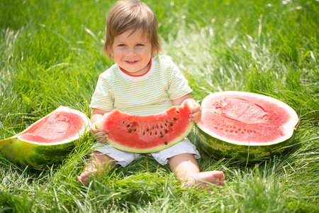Happy child with big red slice of watermelon sitting on green grass. Healthy eating conceptの写真素材