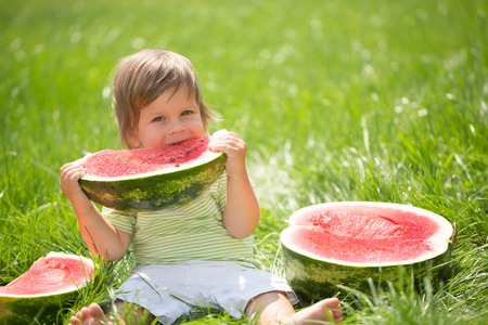 Happy child with big red slice of watermelon sitting on green grass. Healthy eating conceptの写真素材