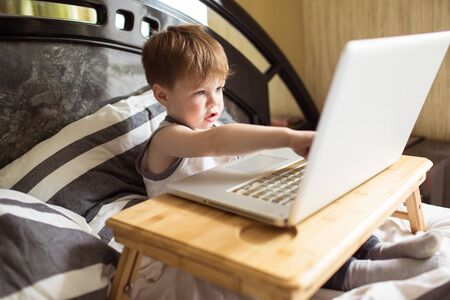 Toddler boy laying on the bed and loooking to pc at home, indoors. Child with computerの写真素材