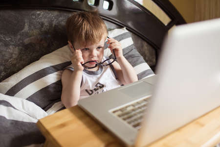 Toddler boy in glasses laying on the bed and loooking to pc at home, indoors. Child with computerの写真素材