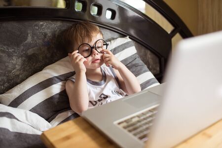 Toddler boy in glasses laying on the bed and loooking to pc at home, indoors. Child with computerの写真素材