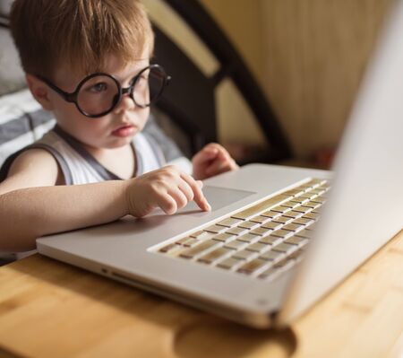 Toddler boy in glasses laying on the bed and loooking to pc at home, indoors. Child with computerの写真素材
