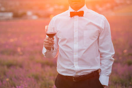 Elegant young guy drinking wine on lavender field at sunset timeの写真素材