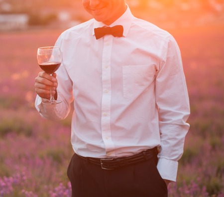 Elegant young guy drinking wine on lavender field at sunset timeの写真素材