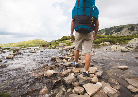 Hiker man with backpack crossing a river on stones in bulgarian mountains. Hiking and leisure theme. Image with sunlight effectの写真素材