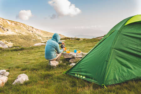 Young man drinking tea at sunrise in mountains. Mountain camping concept.の写真素材