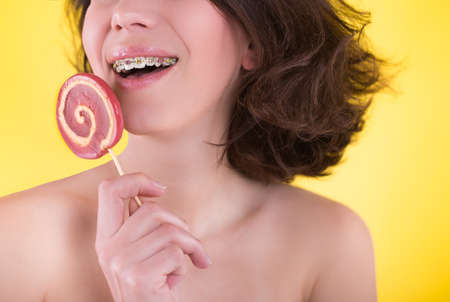 Close-up shot of girl with multicolored bracket systems and lollipop on yellow background