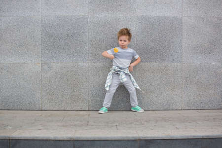 Fashion baby boy in gray wear and mint sneakers stands on a gray wall background. Trendy boy with lollipop standing on the street.の写真素材