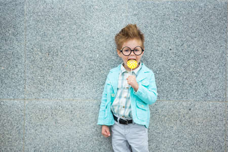 Fashion baby boy in mint jacket stands on a gray wall background. Trendy boy with lollipop standing on the street.の写真素材