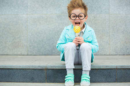 Fashion baby boy in mint jacket and sneakers stands on a gray wall background. Trendy boy with lollipop standing on the street.の写真素材