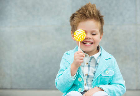 Fashion baby boy in mint jacket and sneakers stands on a gray wall background. Trendy boy with lollipop standing on the street.の写真素材