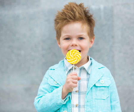 Fashion baby boy in mint jacket and sneakers stands on a gray wall background. Trendy boy with lollipop standing on the street.の写真素材
